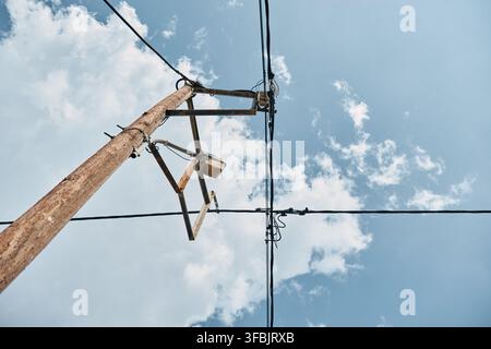 Vue en contre-plongée d'un poteau utilitaire en bois érodé avec des lignes électriques noires s'étendant sur un ciel bleu vif rempli de nuages blancs. Banque D'Images