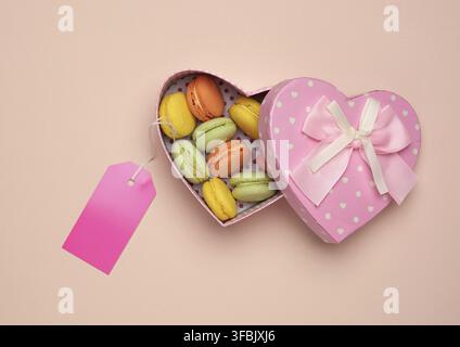 des macarons multicolores cuits ronds sont disposés dans une boîte en carton rose en forme de coeur sur un fond beige, vue du dessus Banque D'Images