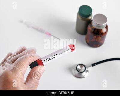Main gantée tenant un tube d'échantillon de sang étiqueté Test ADN avec des équipements médicaux et des flacons de pilules sur fond blanc Banque D'Images