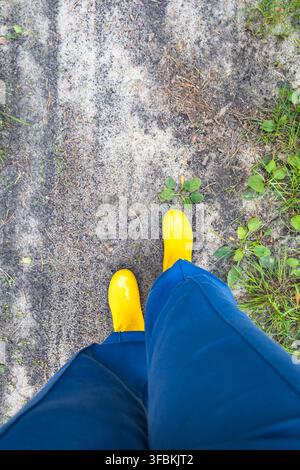 Vue de dessus à la première personne d'une personne portant des bottes en caoutchouc jaune vif et un pantalon bleu marchant le long d'un chemin de terre sablonneux dans un parc en plein air. Banque D'Images