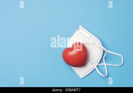 cœur rouge se trouve sur un masque médical blanc jetable, fond bleu, espace de copie Banque D'Images