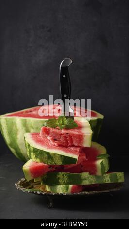 Pile de tranches de pastèque rouge mûre dans une assiette en métal, plantée dans un grand couteau. Fond noir Banque D'Images
