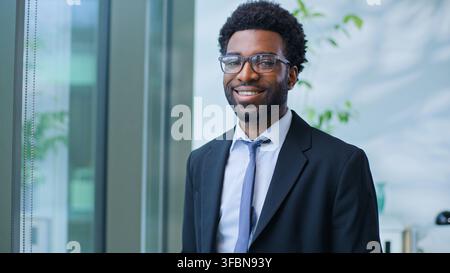 Close-up d'un homme d'affaires afro-américain joyeux et enthousiaste souriant et regardant l'appareil photo, homme d'affaires, type masculin, professionnel près d'une fenêtre, agent, sourire, pose Banque D'Images