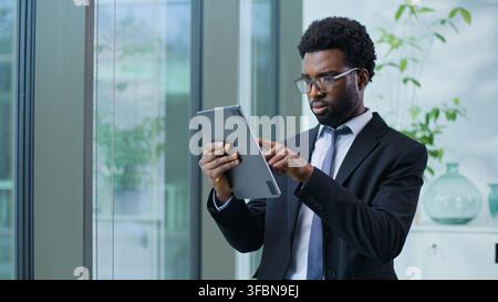 Homme d'affaires afro-américain sérieux concentré à parcourir une tablette numérique, travaillant avec le travail en ligne, homme d'affaires ethnique, type masculin avec la technologie Banque D'Images