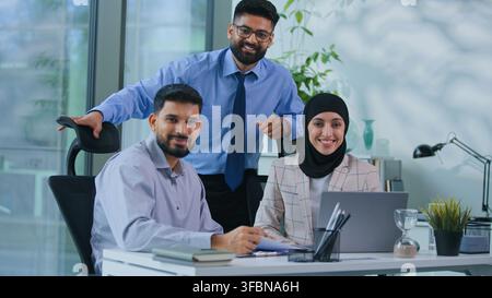 Équipe d'affaires musulmane : trois hommes d'affaires arabes souriant à table, satisfaits des bons résultats de travail avec un ordinateur au bureau, des collègues indiens travaillent Banque D'Images