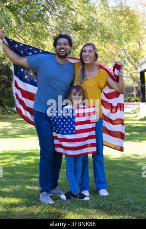 Famille diversifiée debout sur la pelouse tenant un grand drapeau des États-Unis avec un enfant agitant un petit drapeau Banque D'Images