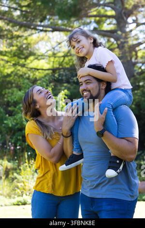 Famille posant dans le parc sous les arbres, père portant sa fille sur ses épaules, maman riant, baskets, montre. Trio, en plein air, ensoleillé, canopée, verdure, fleurs Banque D'Images
