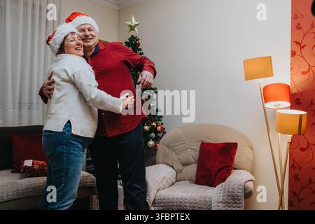Couple de personnes âgées portant des chapeaux de Noël et s'embrassant près d'un sapin de Noël dans une scène de vacances chaleureuse dans un salon Banque D'Images