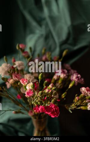 Close-up d'un bouquet d'œillets roses et blancs dans un petit vase en verre. Les fleurs sont posées sur un fond textile drapé vert foncé avec ambiance lumineuse Banque D'Images