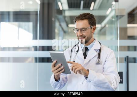 Jeune médecin masculin en blouse blanche et stéthoscope souriant tout en interagissant avec une tablette numérique dans un cadre de clinique ou d'hôpital moderne, représentant l'innovation en médecine et des soins professionnels Banque D'Images