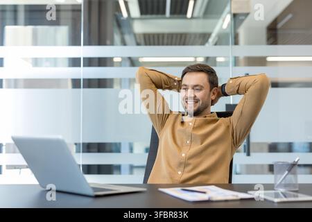 Homme d'affaires souriant penché en arrière dans son fauteuil de bureau, les mains derrière la tête, profitant d'un moment de détente et de satisfaction après avoir atteint ses objectifs et accompli ses tâches Banque D'Images