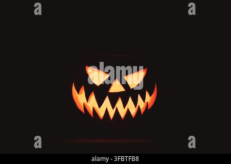 Visage de jack-o'-lanterne lumineux émergeant de l'obscurité avec des dents acérées et une expression maléfique. Fond d'Halloween mettant en vedette une citrouille illuminée et un espace négatif noir dramatique Banque D'Images