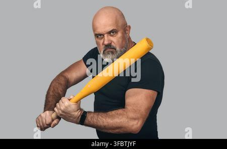 Homme fort barbu tenant une batte de baseball dans une pose intimidante. Fond gris, isolé. Agression, violence, menace. Banque D'Images
