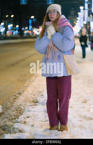 Jeune femme emmitouflée dans un chapeau, une écharpe et des gants attend seule à un arrêt de bus urbain enneigé après le coucher du soleil, debout sur un trottoir glacé sous des lampadaires flous dans un air glacial d'hiver Banque D'Images