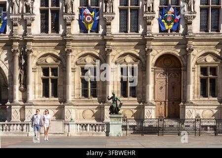Couple passant devant la façade ornée de l'Hôtel de Ville (mairie) décorée de drapeaux cérémoniels, Paris, France. Banque D'Images
