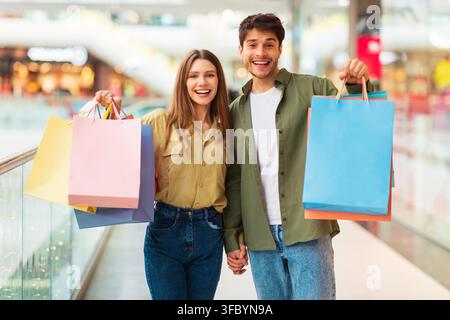 Jeun couple heureux tenant des sacs de courses dans un centre commercial, expression joyeuse Banque D'Images