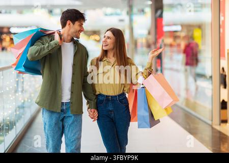 Jeun couple heureux d'origine mixte faisant ses courses ensemble dans un centre commercial, tenant des sacs de courses colorés Banque D'Images