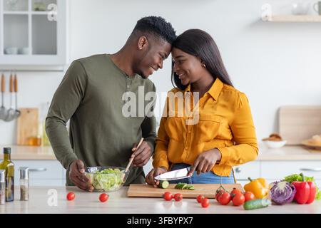 Couple heureux cuisinant ensemble dans la cuisine, préparant une salade fraîche, partenaires multiculturels Banque D'Images