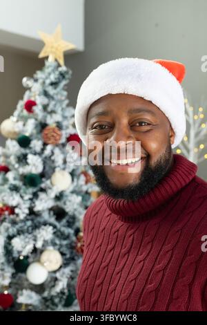 Homme africain souriant à l'objectif à la maison portant un bonnet du Père Noël, un pull bordeaux à côté d'un arbre, espace de copie Banque D'Images