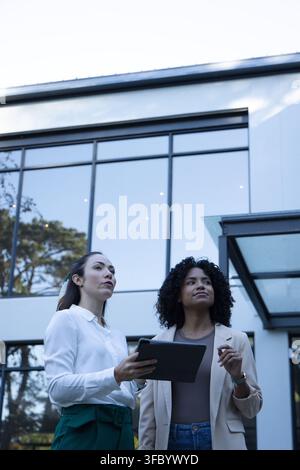 Femmes d'affaires diverses inspectant la façade vitrée et la canopée métallique à l'extérieur du bureau, tenant une tablette Banque D'Images
