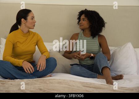 Diverses amies féminines discutant et assises sur un lit portant des hauts moutarde et rayés, télécommande Banque D'Images