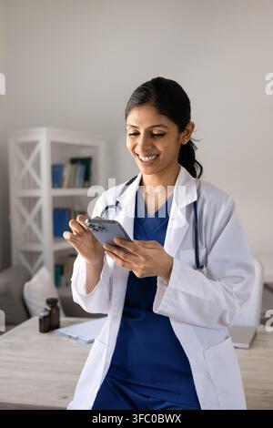 Médecine indienne souriante debout sur son lieu de travail utilisant un smartphone Banque D'Images