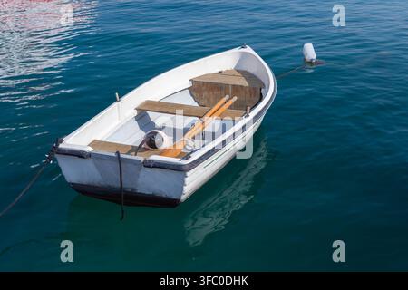 Un petit bateau à rames blanc amarré à une bouée et flottant sur l'eau bleue et calme du port, avec des rames en bois à l'intérieur. Scène paisible et estivale évoquant la détente Banque D'Images