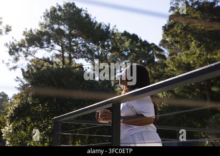 Femme africaine adulte sur un balcon à rambarde métallique, vêtue de vêtements clairs, tenant une tasse et regardant vers le haut, espace de copie Banque D'Images