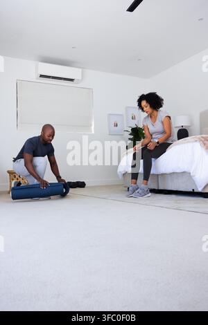 Couple afro-américain en vêtements de sport déroulant un tapis de yoga sombre dans la chambre, posant des haltères Banque D'Images