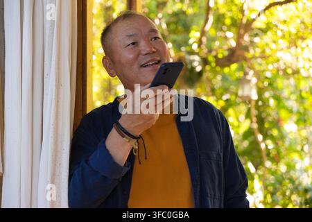 Homme asiatique âgé debout dans l'encadrement de la porte tenant un smartphone à la bouche, portant des bracelets, dans un jardin baigné de soleil Banque D'Images