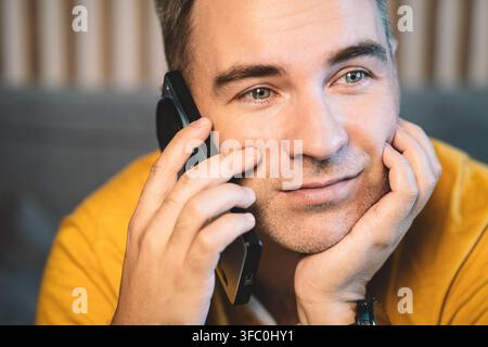 Close-up d'un homme souriant parlant au téléphone, reposant son menton sur sa main. Appel agréable, conversation amicale et atmosphère domestique détendue Banque D'Images