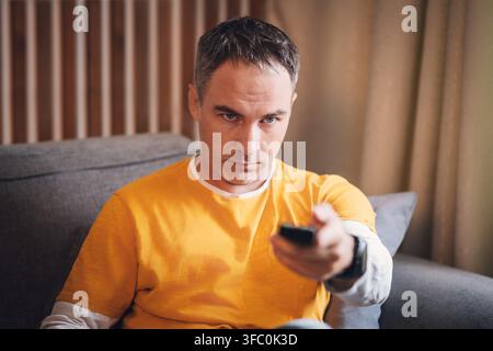 Homme d'âge moyen en t-shirt jaune moutarde assis sur un canapé gris tenant une télécommande TV ; expression concentrée et sérieuse. Lattes en bois et rideaux beiges i Banque D'Images