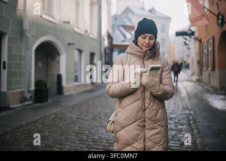 Une femme se tient sur une rue pavée et regarde son téléphone tout en portant des vêtements chauds par une journée fraîche. Banque D'Images