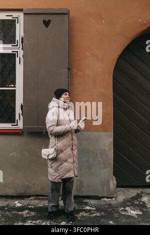 Une femme est à l'extérieur d'un bâtiment en hiver. Elle porte un manteau chaud et tient un téléphone tout en regardant autour d'elle. Banque D'Images