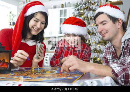 Famille traditionnelle heureuse composée de papa, maman et fille portant des chapeaux de Père Noël festifs, jouant à un jeu de société à table dans une cuisine blanche avec un sapin de Noël et des décorations. N Banque D'Images