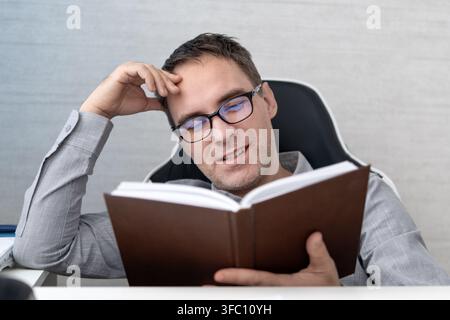 Homme souriant lisant un livre sur une chaise de bureau, profitant de l'apprentissage, du développement professionnel ou de la croissance personnelle pendant une pause de travail relaxante. Banque D'Images