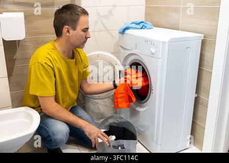 Un homme portant un t-shirt jaune charge un vêtement orange dans une machine à laver ; une serviette bleue repose dessus, un panier à linge se trouve à proximité, dans un carrelage compact Banque D'Images