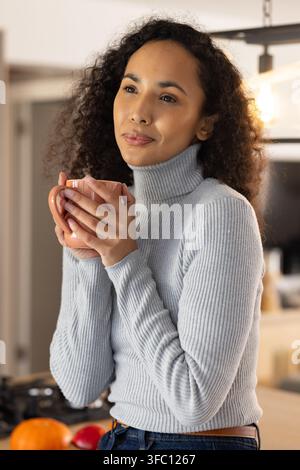 Femme afro-américaine portant un pull gris clair tenant une tasse et regardant dans la cuisine de la maison Banque D'Images