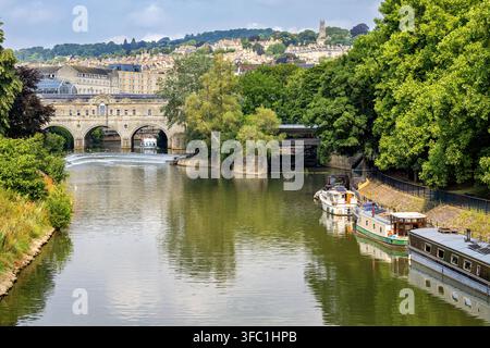 Pont historique de Pulteney avec un barrage dans la rivière Avon par une journée d'été ensoleillée, entouré de narrowboats et d'arbres verts à Bath, Somerset, Angleterre. Banque D'Images