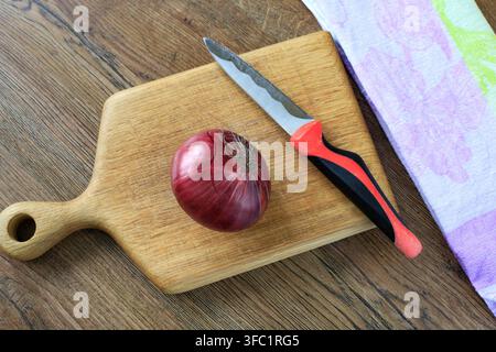 Tête d'oignon violet non pelée posée sur une planche à découper en bois naturel avec un couteau bien aiguisé sur une table rustique. Concept de préparation des repas, de produits bruts et de guérison Banque D'Images