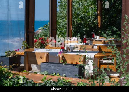 Une charmante terrasse de restaurant en plein air avec des tables en bois dressées et une végétation luxuriante, offrant une vue paisible sur l'océan bleu vif par une journée ensoleillée Banque D'Images