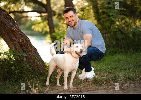 Homme d'âge moyen caressant son chien souriant dans un parc vert près d'un lac Banque D'Images