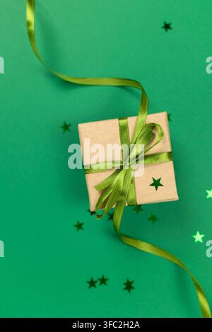 Boîte cadeau enveloppée dans du papier kraft brun avec un nœud en ruban de satin vert et des confettis en forme d'étoile sur fond vert Banque D'Images