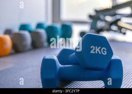 Vue rapprochée de deux haltères bleus de 2 kg reposant sur un tapis de gym texturé, avec une sélection d'autres poids flous en arrière-plan Banque D'Images