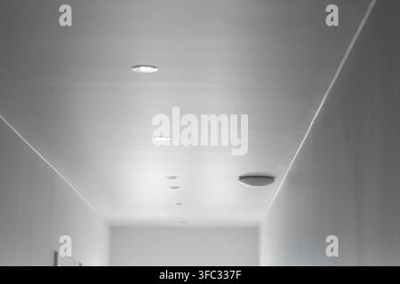 Un couloir intérieur minimaliste présente un plafond blanc immaculé avec plusieurs spots encastrés et un dispositif d'alarme fumée circulaire monté au plafond Banque D'Images