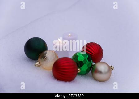 Close up de baubles colorées variées et de deux bougies lavande brûlantes reposant sur un lit de neige blanche. Décorations de fête détaillées avec des couleurs vives a Banque D'Images