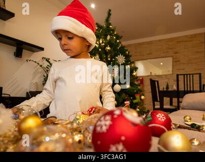 Enfant portant un chapeau rouge contemple des ornements festifs. Des boules dorées et rouges l'entourent. Les lumières de l'arbre scintillent doucement en arrière-plan. Banque D'Images