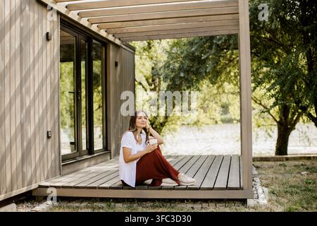 Une jeune femme se détend sur une terrasse en bois, sirote son café tout en profitant de la vue sereine sur la rivière entourée de verdure. Banque D'Images