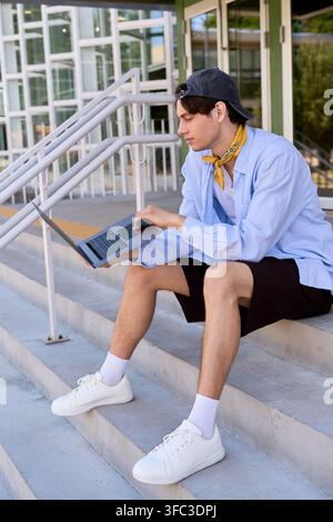 Un étudiant en chemise bleue et short utilise un ordinateur portable assis sur les marches du campus Banque D'Images