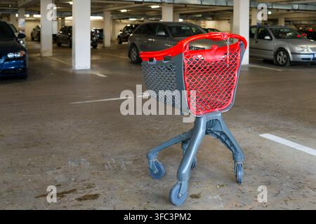Un panier d'achat rouge et gris vide se trouve dans un parking intérieur entouré de voitures garées, illustrant les achats de détail et la vie des consommateurs Banque D'Images